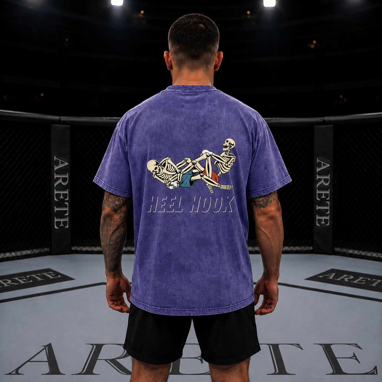 A man viewed from behind wearing a purple t-shirt with a large graphic of two skeletons and the text 'HEEL HOOK' on the back, standing inside an ARETE boxing ring.